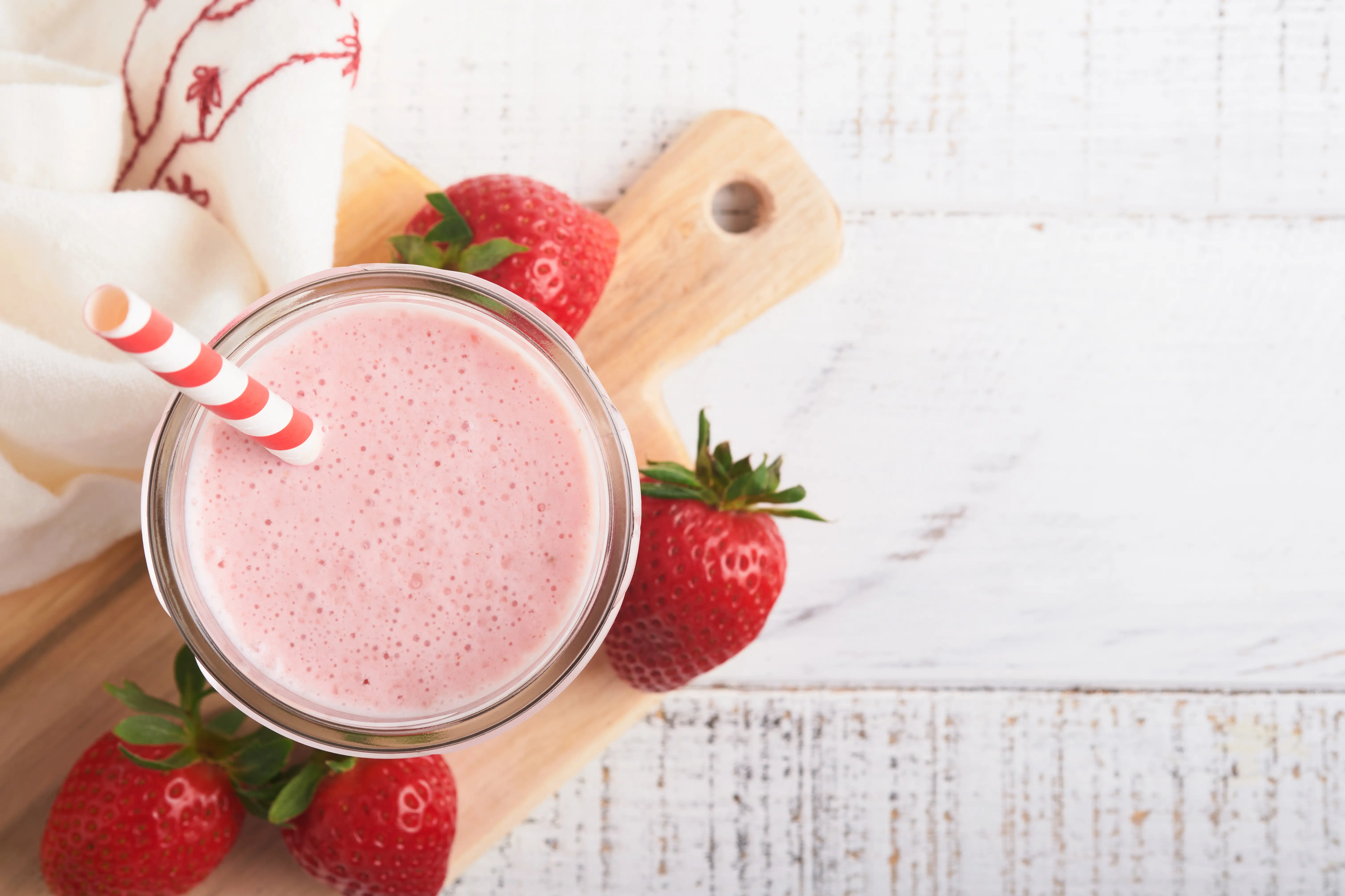 a glass of pink smoothie with straw and strawberries on a wooden board